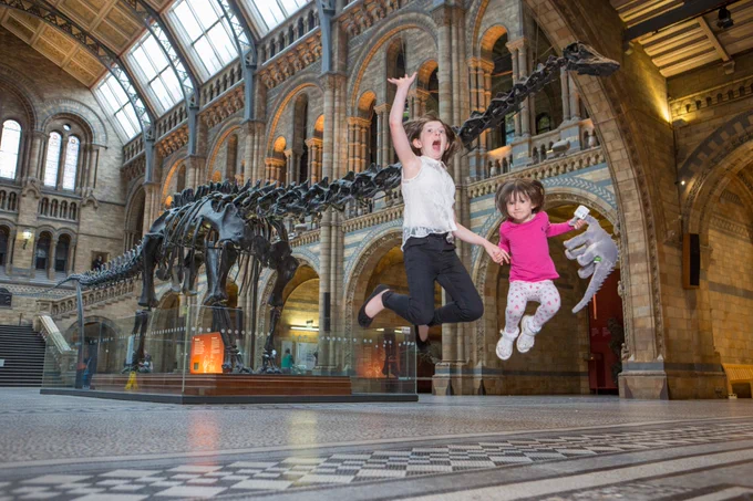 Two girls jumping into the air in front of Dippy at the Natural History Museum. One of them is holding a dinosaur soft toy.