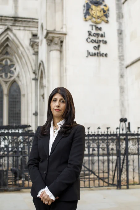 Photo of Lubna Shuja standing outside the Royal Courts of Justice. Asian woman with brown hair, wearing a black blazer and white shirt.