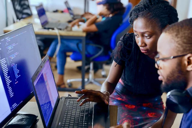 A man with glasses looks at his computer screen as a woman standing next to him  types on his keyboard. Desks with computers and people in the backgound