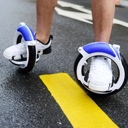 A pair and legs and feet perched on platforms in the center of two hubless wheels balanced on some wet asphalt with yellow and white road markings. The wheels have a white plastic shroud holding the bearings and foot platforms (pedals?) and a blue rubberizsed grip on the top.