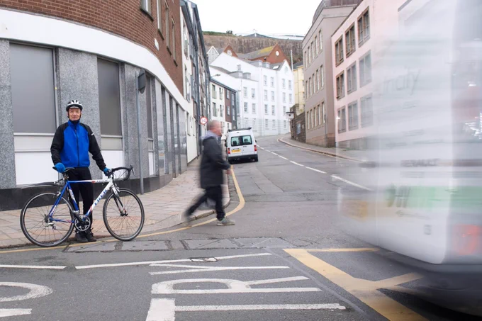 A man in sporting attire stands with a racing bike on a street in Saint Helier, Jersey. A bus passes in the foreground.