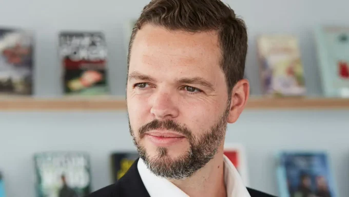 Tim Whiting smiling, looking off-camera. Blurred in the background is a white wall with bookshelves and books face-out on display.