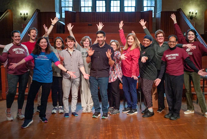 A group of 13 people standing together for a posed photo. Their hands are in the air and are smiling.