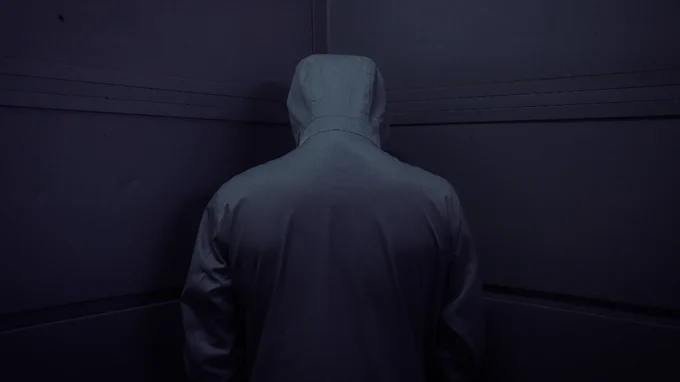 An image of a hacker like persona draped in a dark hoodie facing a corner their back to the camera. Image credit: De an Sun on Unsplash.