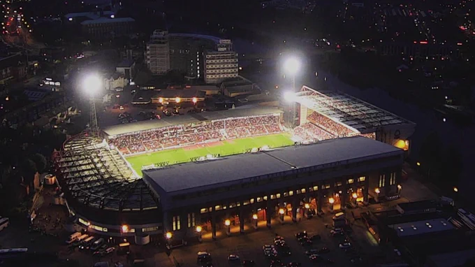 Aerial photo of the City Ground in Nottingham, taken from a drone. The stadium is illuminated by the floodlights amid the backdrop of the dark, night sky.