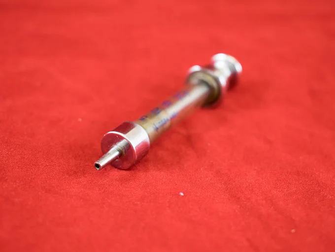 A small metal and glass syring lying on a piece of red cloth. The focus of the camera is on the tip of the syringe. There is blue lettering on the glass of the syringe to measure dosage. 