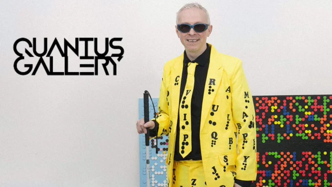 Clarke stood proudly beside his colourful and tactile Braille art. He is holding a cane and wearing a yellow suit covered in the Braille alphabet.