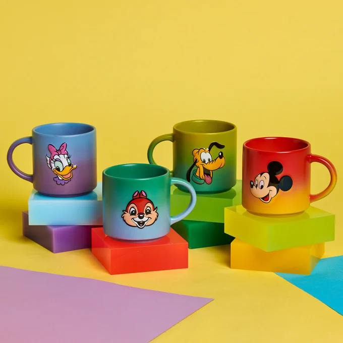 Photo of character essential mugs on blocks. From left to right mugs are: Blue and Purple featuring Daisy, Blue and Green featuring Chip, Green featuring Pluto, and Red and Orange featuring Mickey.