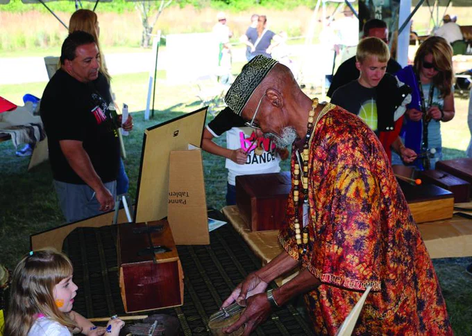 An older Black man wearing a brightly colored traditional African outfit shows a drum to a young child. 