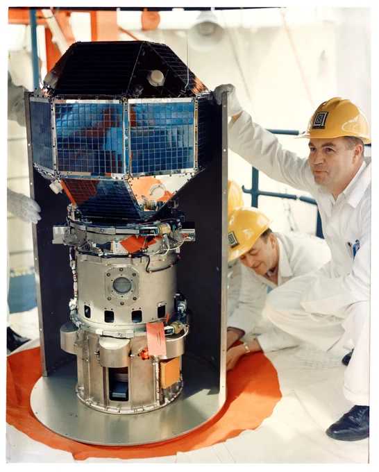 Two employees in white jumpsuits and yellow hard hats working on the military satellite communications program.