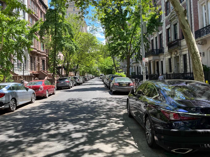 View looking down a city street with leafy trees overhead and parked cars on each side of the street obscuring the view of the narrow sidewalk.