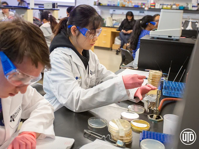 Two UT Dallas students wearing white lab coats and protective glasses conduct lab work