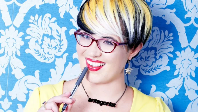 Sarah Rees Brennan smiling at the camera. She is wearing a yellow top and red glasses and is in front of a blue and white floral wall.