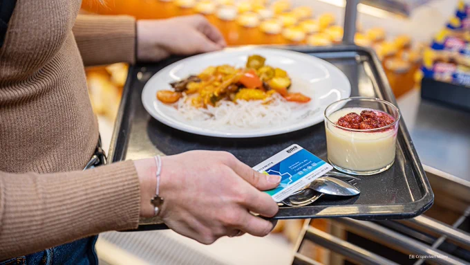 Detailfoto eines Tabletts, auf dem sich ein Teller mit Essen sowie ein Schälchen mit Nachtisch befindet. Im Bild sind auch noch die Hände der Person abgebildet, die das Tablett trägt. In der rechten Hand hält die Person zudem noch die Campuscard der TU Dresden.