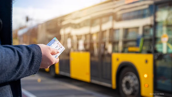 Fotoaufnahme einer Hand, die die Campuscard, den digitalen Studierendenausweis der TU Dresden, hält. Im Hintergund ist leicht unscharf ein gelber Bus abgebildet.