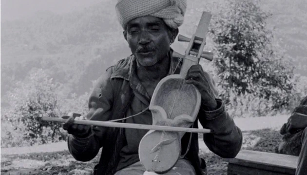 A black and white photograph of a man sat down wearing a light coloured turban and dark clothing. He is playing a traditional strings instrument, behind him are trees.