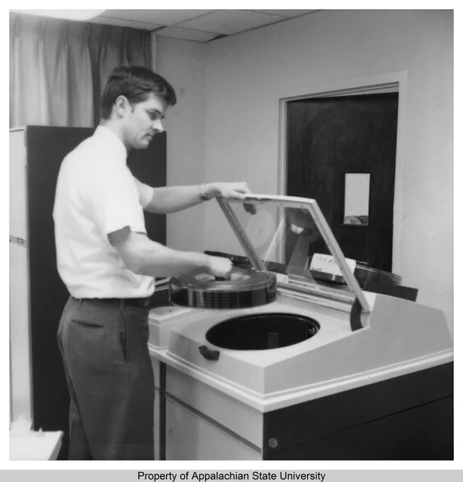 Description: This image shows the Computer Center at Appalachian State University (1967-current), circa 1970. The Center began in 1968 and was primarily used as a laboratory for the computer science courses, and for aiding University administrative offices. This image shows an employee loading a disk pack into a direct access storage subsystem. By 1974, Appalachian State was operating both an IBM 1130, introduced 1965, and a UNIVAC 9400, introduced 1969. Both IBM-compatible systems included disk drive units using a disk pack, a collection of platters or "heads" for storing data, with a maximum capacity of 7.2 megabytes. It was mounted on a common actuator which would read or write information to the disks. By 1976, they were operating UNIVAC Series 70/46 and 90/60