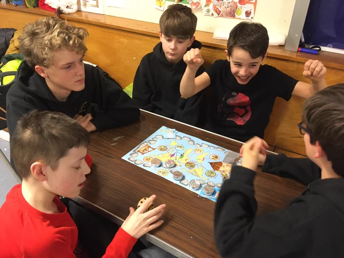 5boys sitting around a table on which is placed a board game. One boy has both hands in the air, miming a spider