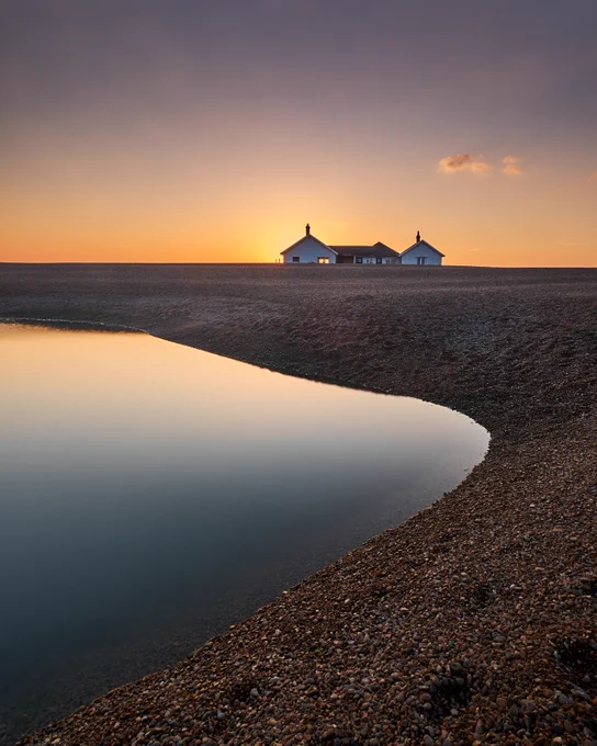The Sun setting behind a house standing alone on a shingle beach, with a calm pool of water reflecting the sky in the foreground.