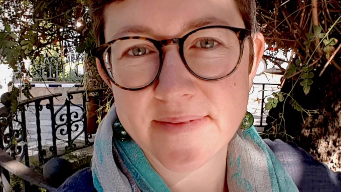Close-up of Rebecca Wragg Sykes smiling at the camera, wearing round glasses. In the background is a tree and a circular iron fence.