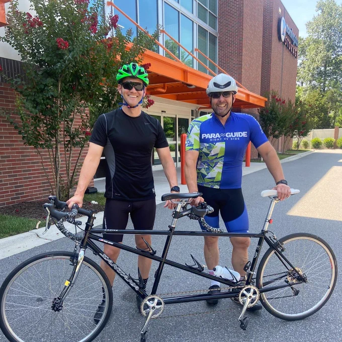 Paul and Jeffrey pose with a tandem outside of Morningstar; both are smiling and wearing cycling gear and helmets