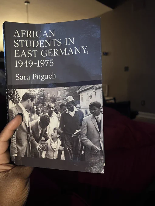 A photo of my hand holding Sara Pugach’s book “African Students in East Germany, 1949-1975”. The cover shows a black and white photo of several African students crowding around a young white boy. The students are smiling. The boy is looking up at a student bending over to talk to him.