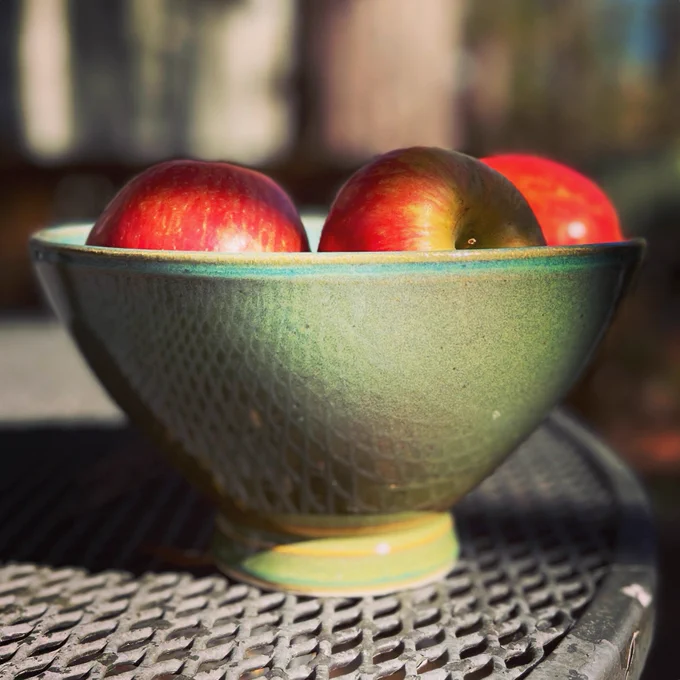 Photo of a green glazed pottery bowl filled with red apples on a black rod iron table outside by Megan Flynn Hopkins of EURUS ceramics in Richmond, Virginia, handmade art for your home, winsome and puckish.