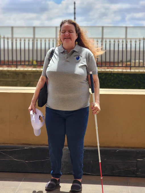 WBU President Martine Abel-Williamson in a grey polo shirt with a WBU logo and a blue jean. Holding a white cane and a white cap on both hands. Smiling at the camera, with her hair blowing in the wind.