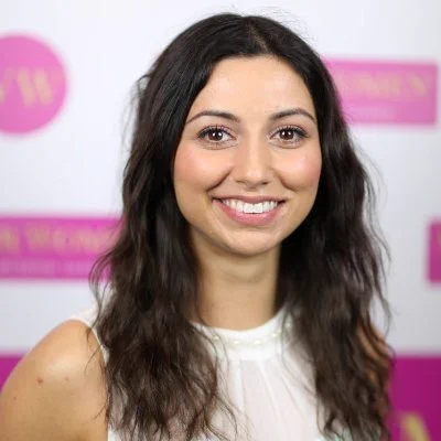 A pic of award winning sound expert Emma Butt who is hosting the Channel 4 event about diversity in TV Post Production. She is standing against a pink and white board, and has long dark hair. She is smiling and is wearing a white sleeveless top. 
