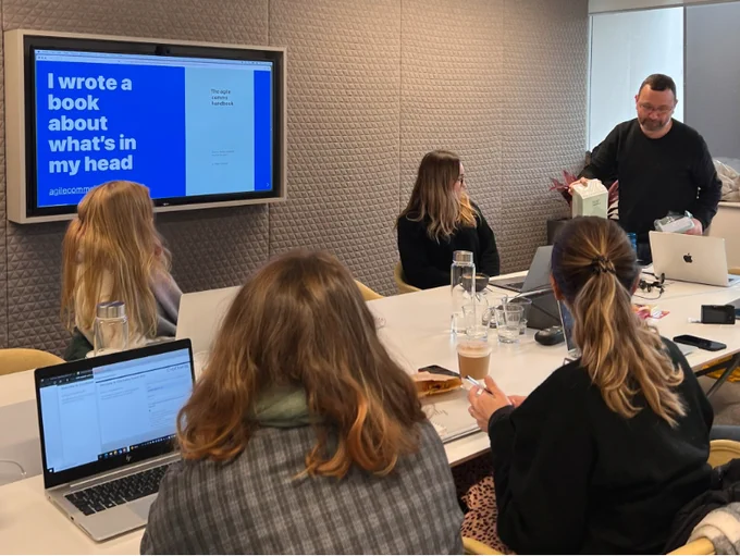 A group of people sat around a table. Behind them is a blue screen with the words 'I wrote a book about what's in my head. The agile comms handbook'