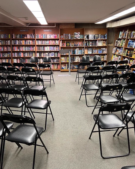 Black chairs are lined up in rows to face a podium. Behind the podium is a wall of bookshelves.
