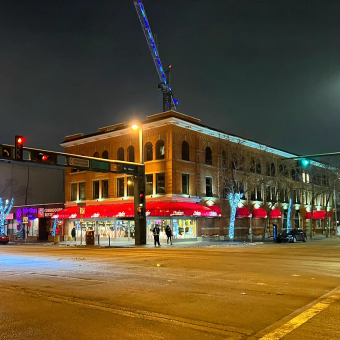 A downtown building is lit up by lamplight and shop lights in the dark. It has a red Audreys sign wrapped around it. In the foreground is a traffic light.