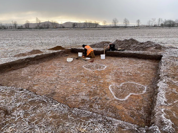 Archaeologist at work in a square trench with frosty field and tree line in background