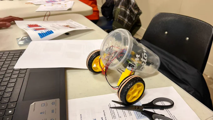 Robotic toy car built using micro:bits and a recycled coffee cup. It’s sitting on a table with instruction papers and a laptop.