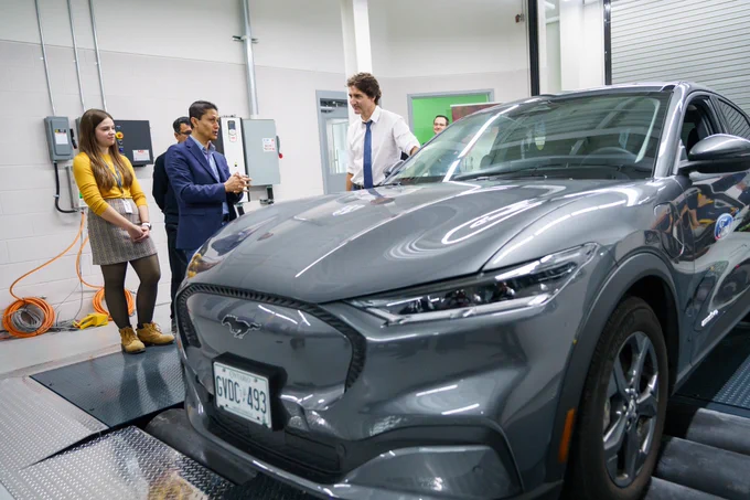 Prime Minister Justin Trudeau and Dr. Narayan Kar are standing indoors and speaking with each other. Two University of Windsor students are standing behind them. In front of them, an electric vehicle is visible.