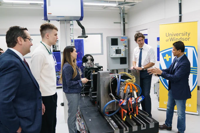Prime Minister Justin Trudeau and Dr. Narayan Kar are standing indoors and speaking with each other. They are standing in front of machinery. Two University of Windsor students and Member of Parliament Irek Kusmierczyk are standing across from them.