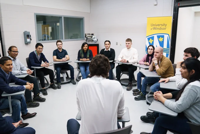 Prime Minister Justin Trudeau is sitting at a desk indoors. University of Windsor students are sitting in a semi-circle in front of him.