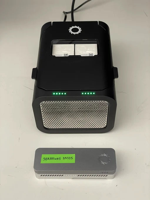 A black brushed metal P2 Solo Nanopore sequencer, the size of a cheese grater, with two positions for attaching PromethION flow cells. A MinION sequencer, size of a USB dongle, is shown in bottom for scale.