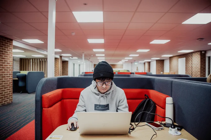 A student is sitting in one of Sussex Library study booths on the first floor. He is wearing a hat and glasses and browsing on his laptop.