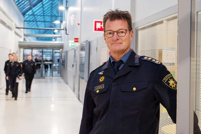 Yliopettaja Matti Honkanen katsoo kameraan seisoen Pelastusopiston käytävällä, opiskelijoita taka-alalla.