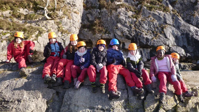 Pupils wearing helmets and waterproofs sat on rocks in the sunshine on the beach