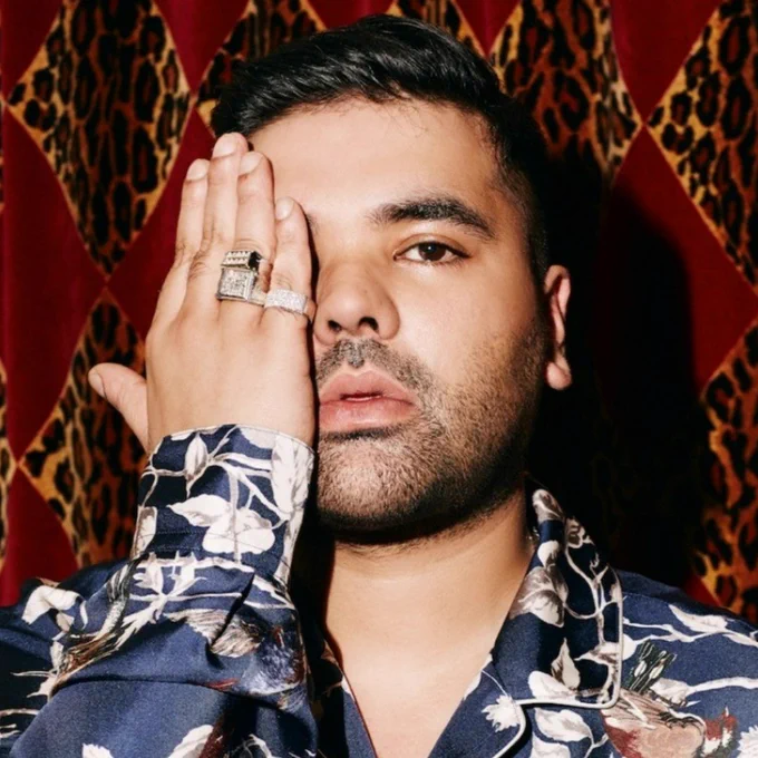 Close-up of Naughty Boy, wearing a flamboyant shirt and holding one hand up to his face.