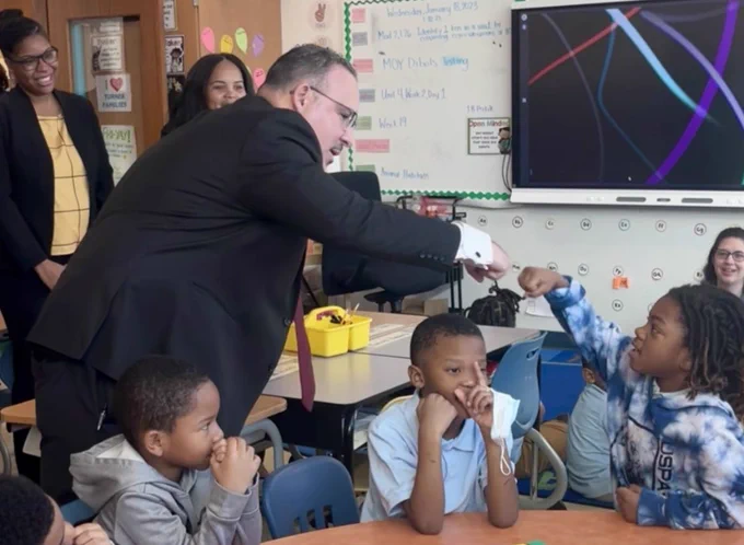 Secretary Cardona fist bumps a student at Turner Elementary in a sunny, engaged classroom with guests and school supplies in the background.