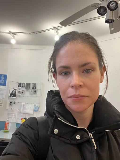 Photo of brown haired woman wearing a winter coat. Over the woman’s right shoulder are two strings of photographs, each photo held with a clothespin, in a cute way - like at a summer camp or daycare.