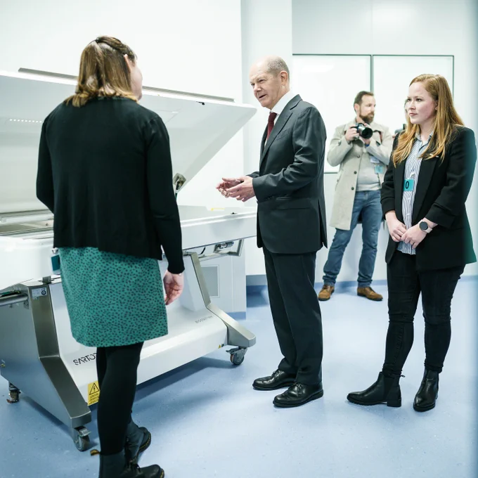Eine Frau in Rückenansicht zeigt Bundeskanzler Scholz eine aufgeklappte Maschine. Eine weitere Frau und ein Fotograf stehen dabei. 