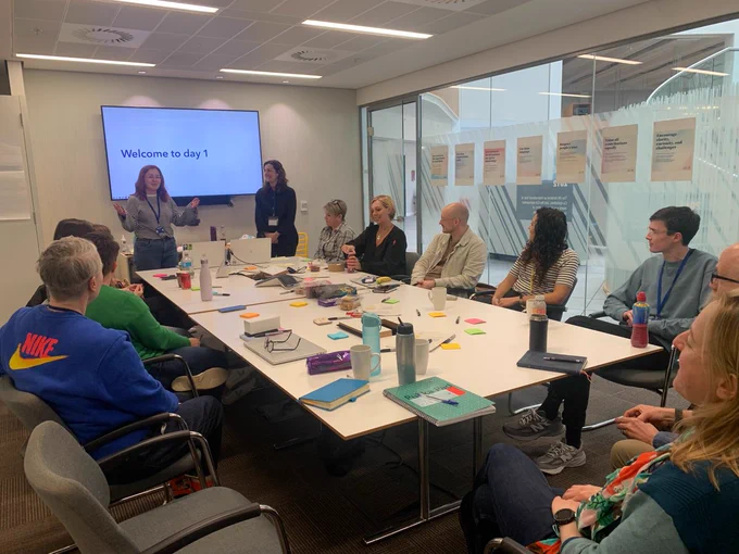 A meeting room with lots of people sat at a table with post-it notes. I am wearing a stripey top and standing next to Marianne, in front of a screen saying 'welcome to day 1'. My arms are half raised and I look like I am doing the shrug emoji.