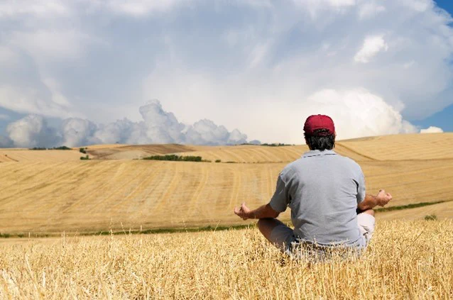 In a golden field of recently harvested grain, a person with their back to the camera is sitting toward the cloud-filled blue sky and looks to be meditating by the position of the hands and fingers. 