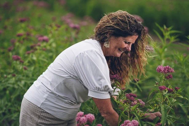 A lady with long hair smiles down as she looks at her flowers growing in the field.