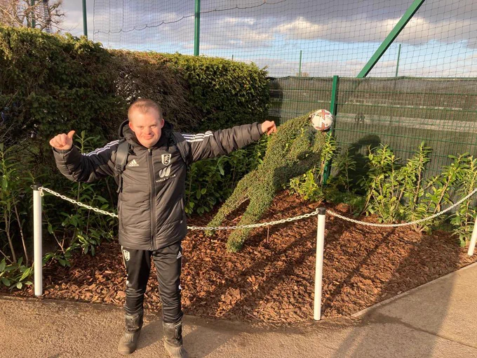 Matthew stands in his FFC Groundsman uniform in front of his newly lanscaped bed. With bushes round perimeter a Goalkeeper shaped topiary bus making a save, wood chippings and a fence around the front