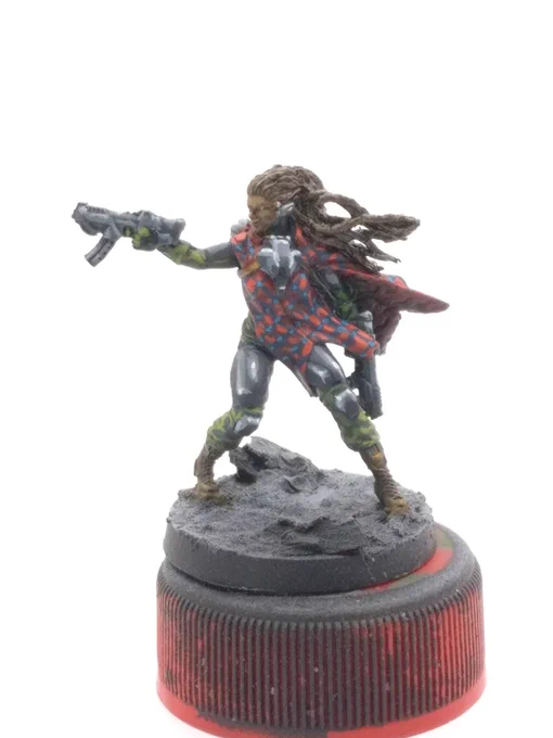 Aida Swanson from Corvus Belli's Infinity Defiance boardgame. humanoid woman with dark skin and braids brandishing a pair of submachineguns. green fatigues, gunmetal armor plates, and a red and blue plaid pattern resembling Maasai shuka cloth.